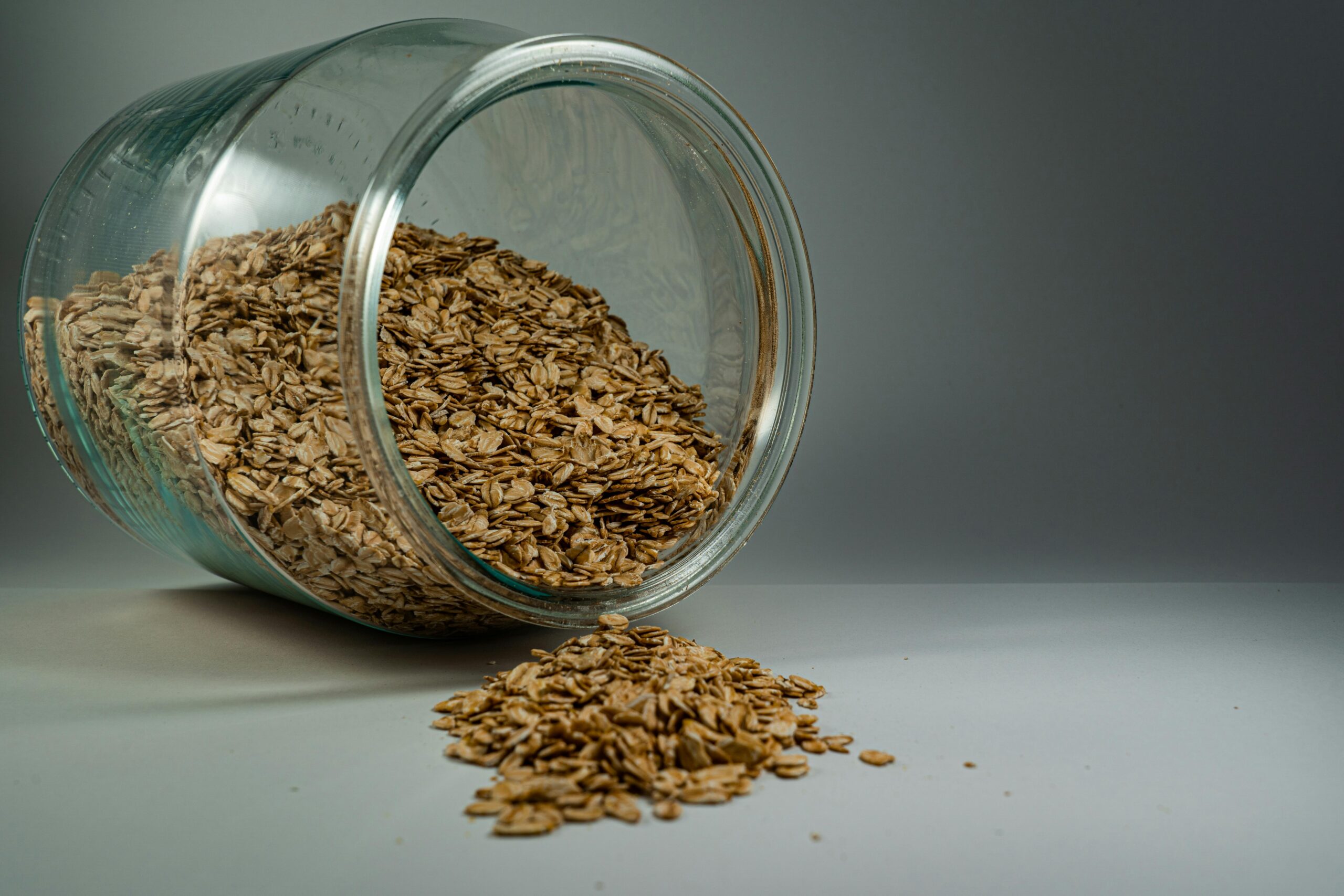 A glass jar spilling rolled oats onto a plain surface, showcasing a rustic breakfast ingredient.